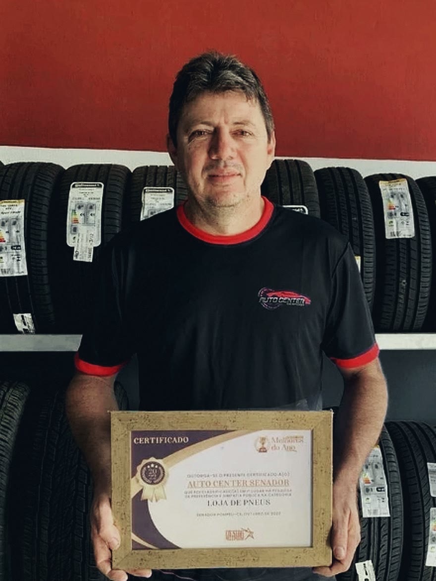 Jonas Bruno, dono da Auto Center, na oficina, em frente à prateleira de pneus, segurando o certificado Melhores do Ano 2022